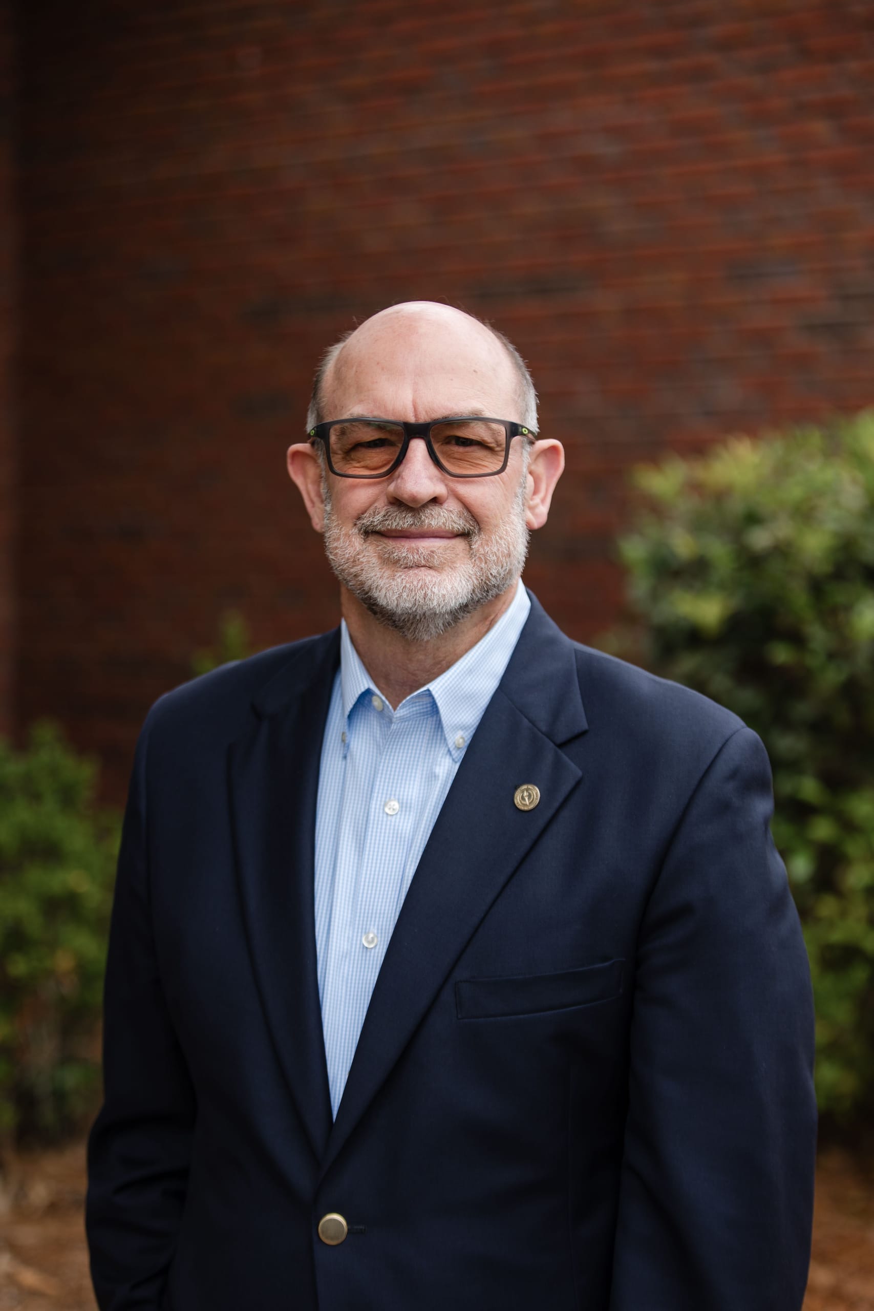 Headshot of a man in glasses and a navy blazer in front of a brick wall